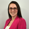 Veronica poses for a headshot, smiling at the camera with long brown hair and glasses, wearing a bright pink blazer over a white top against a plain light background.
