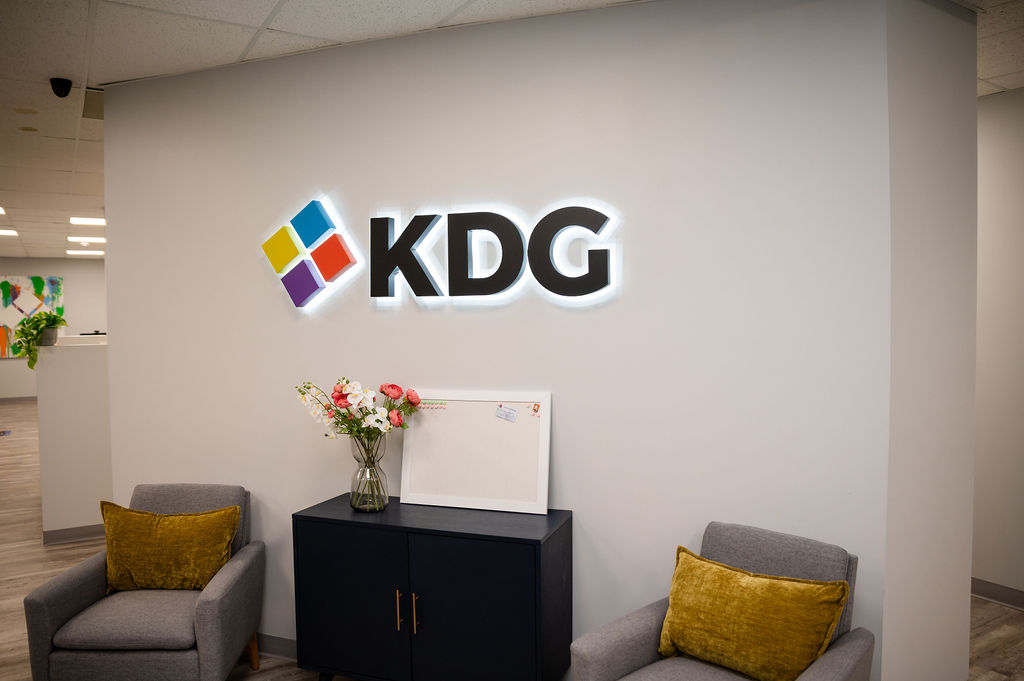 A wall with a lit "KDG" logo, a bouquet of flowers, a blank canvas on a black cabinet, two gray chairs with yellow cushions, and a neatly displayed sitemap.