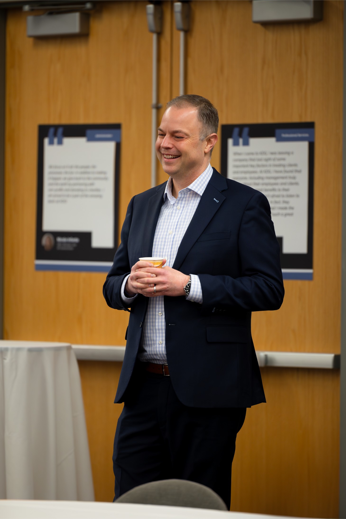A man in a suit holds a cup, smiling, standing in front of wooden doors with posters. A man in a suit holds a cup, smiling, standing in front of wooden doors with posters.