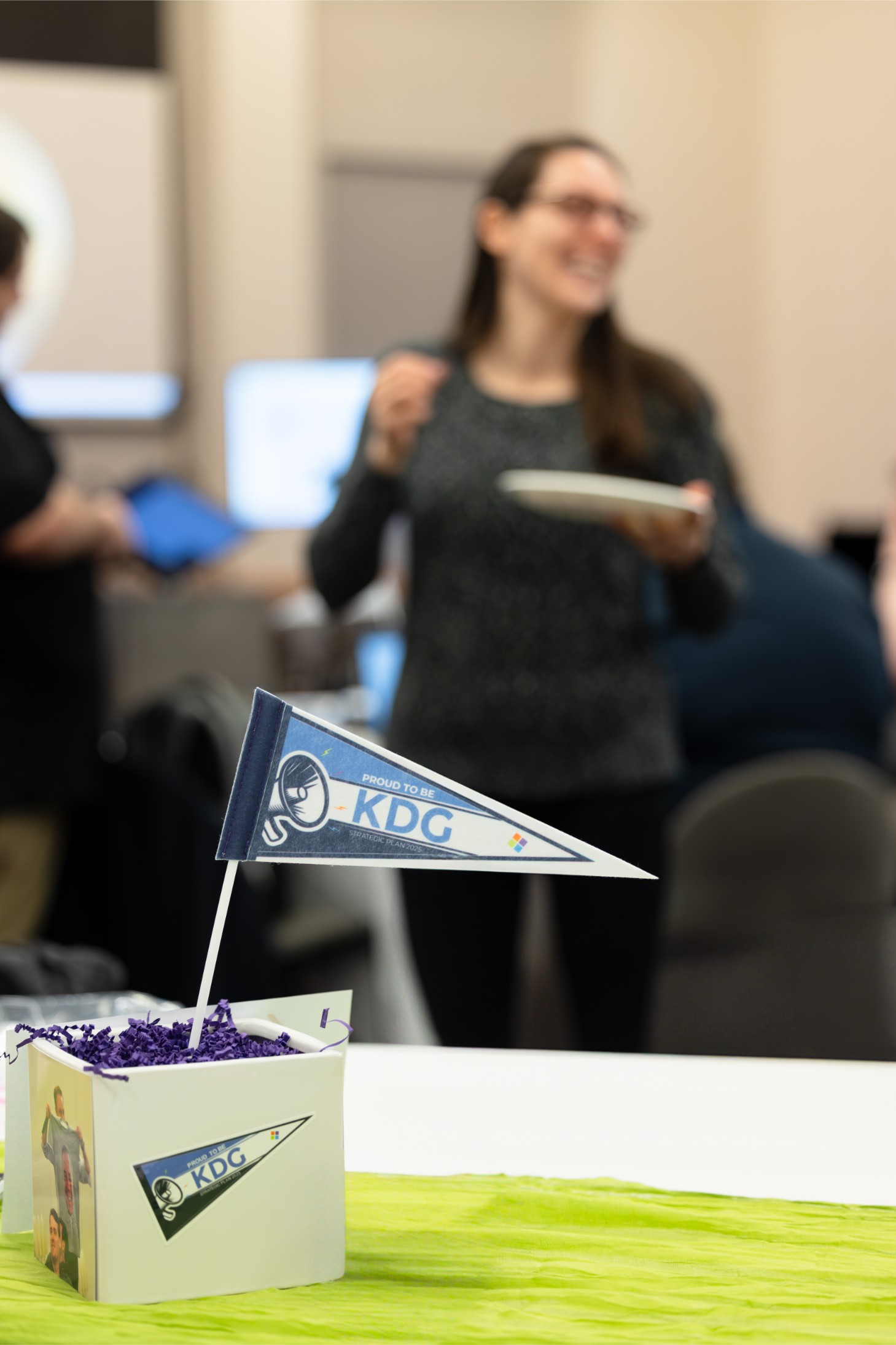 A small flag with “KDG” is on a table centerpiece. A blurred person in the background holds a plate, suggesting a social gathering or event. A small flag with "KDG" is on a table centerpiece. A blurred person in the background holds a plate, suggesting a social gathering or event.