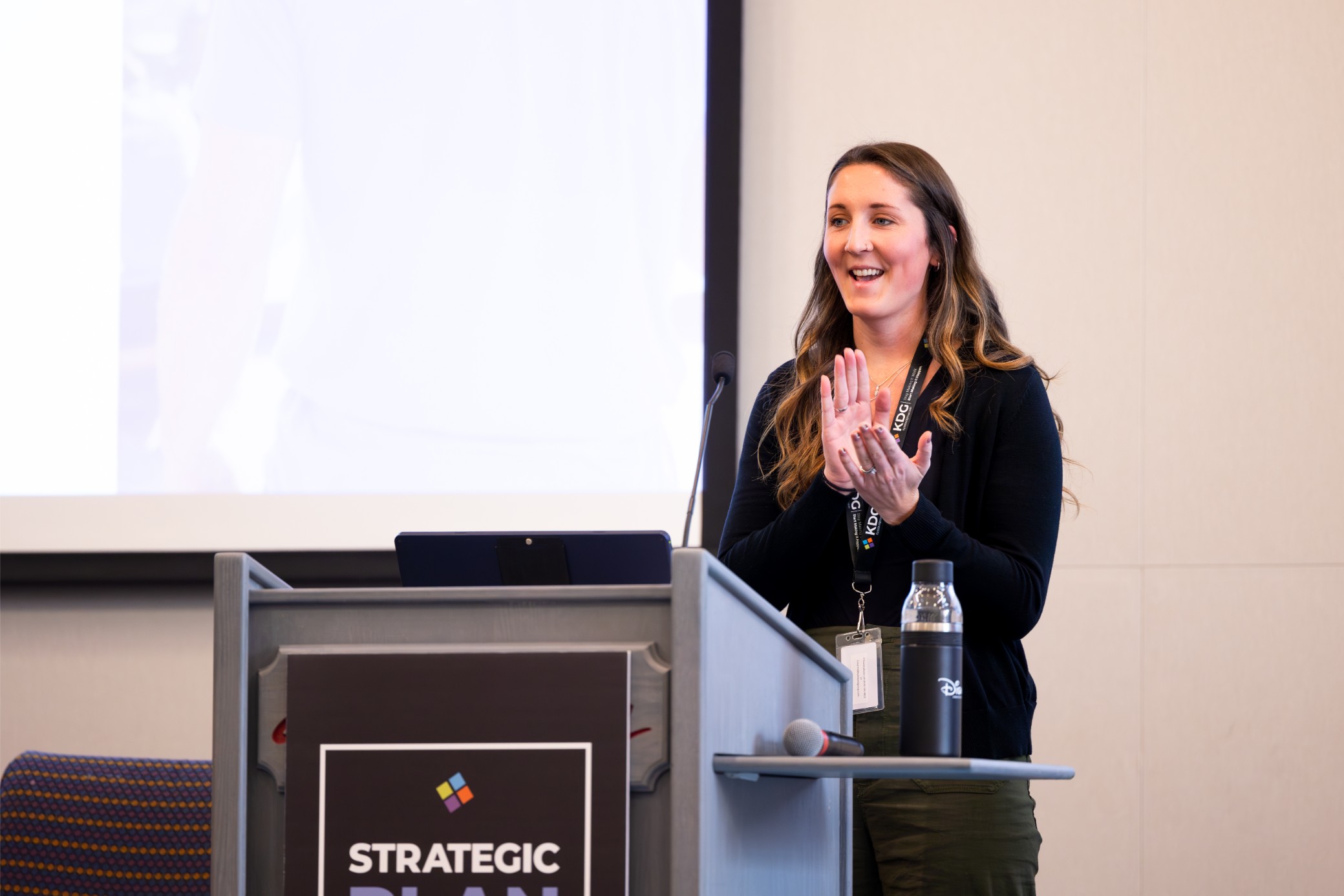 A woman stands at a podium, clapping, during a presentation titled “Strategic Plan.” A water bottle is visible. A woman stands at a podium, clapping, during a presentation titled "Strategic Plan." A water bottle is visible.