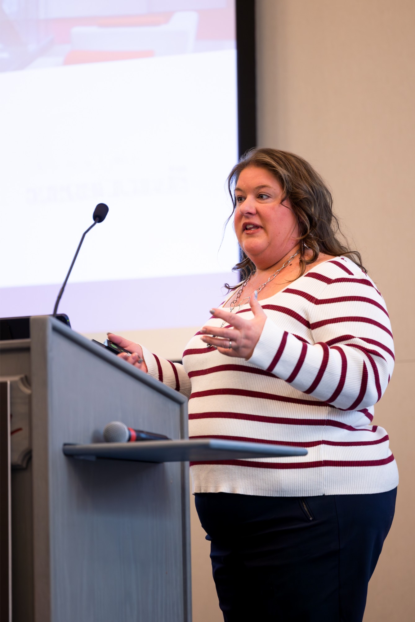 A person in a striped sweater speaks at a podium with a microphone, presenting in front of a screen. A person in a striped sweater speaks at a podium with a microphone, presenting in front of a screen.