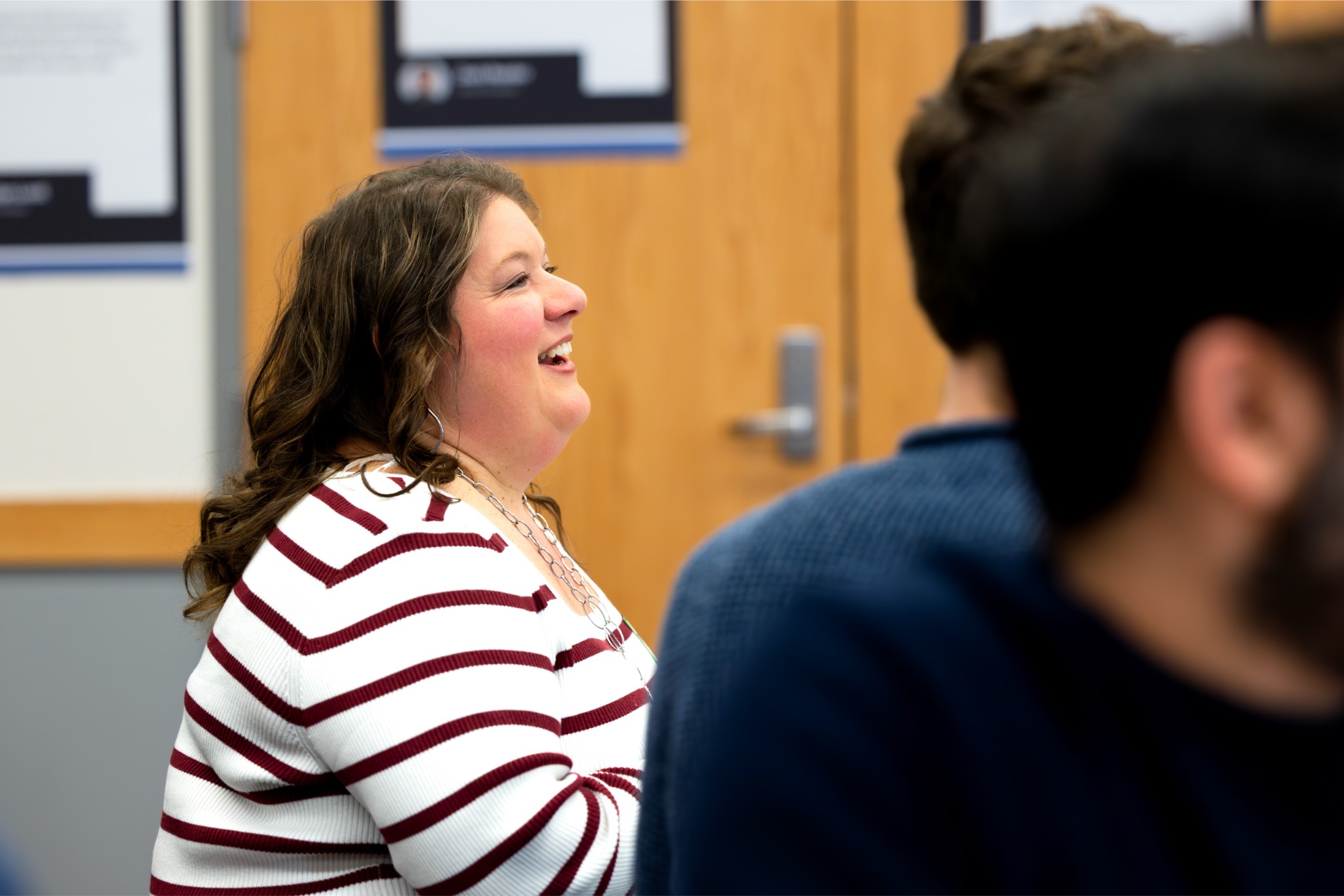 A woman in a striped sweater is smiling and sitting in a room with blurred posters on the wall. A woman in a striped sweater is smiling and sitting in a room with blurred posters on the wall.