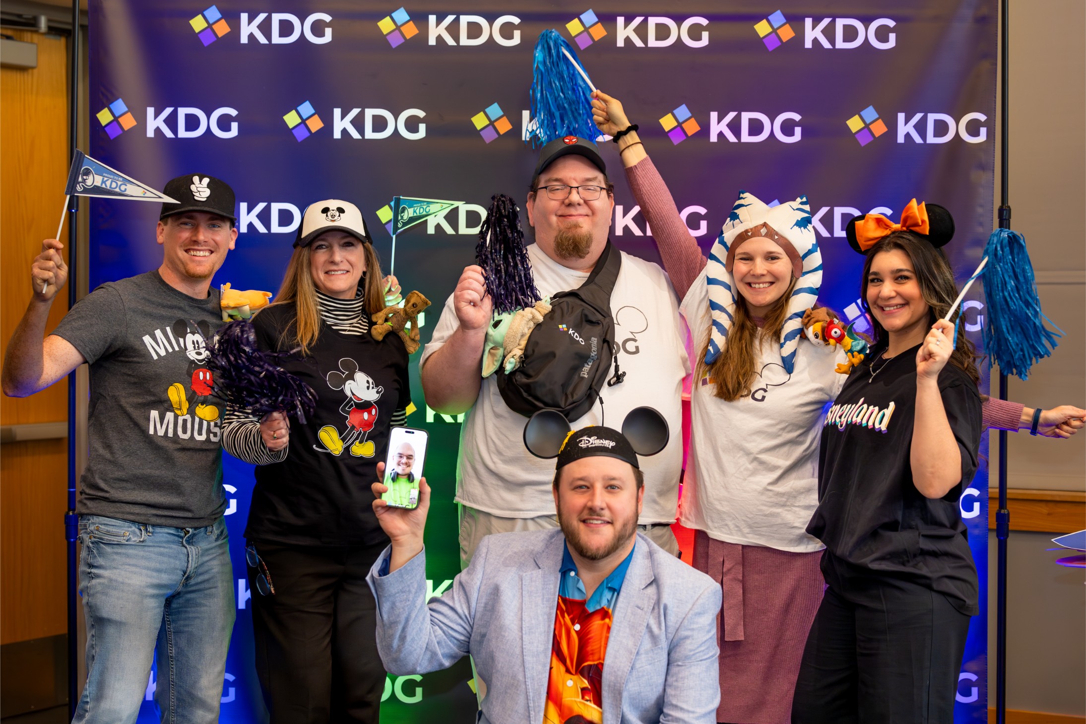 A group of six people pose cheerfully in front of a KDG backdrop, wearing various Disney-themed apparel and accessories. A group of six people pose cheerfully in front of a KDG backdrop, wearing various Disney-themed apparel and accessories.