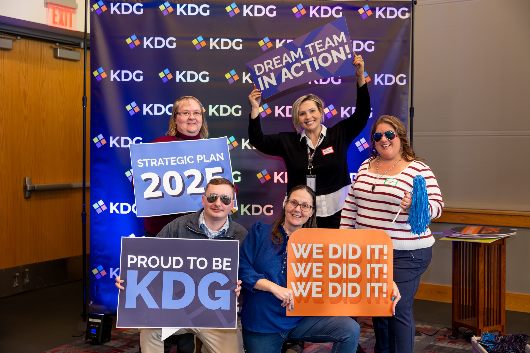 Group of six people holding signs with phrases like "Dream Team in Action" and "Strategic Plan 2025" in front of a KDG backdrop.
