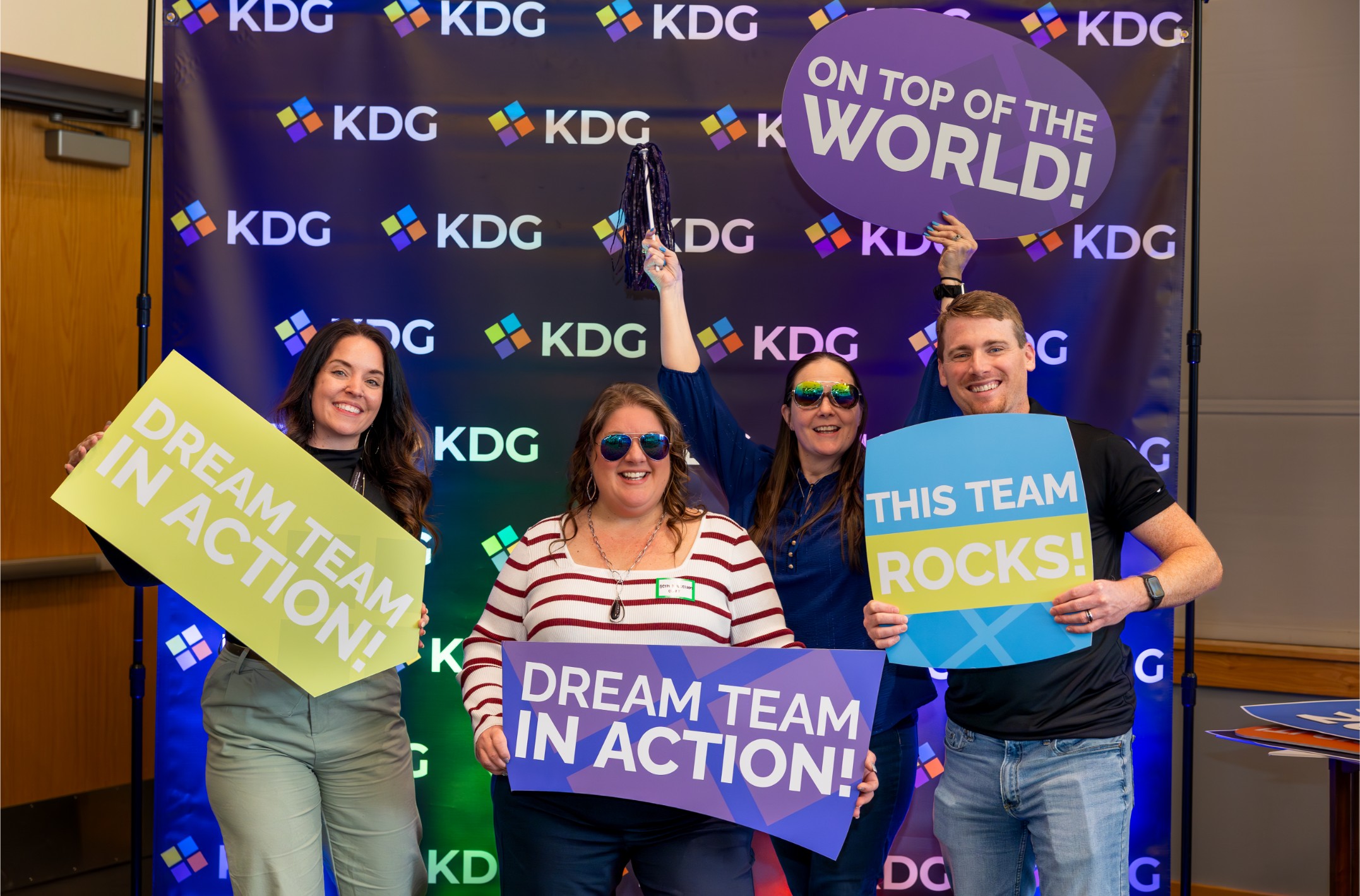Four people holding colorful signs that say “Dream Team in Action,” “On Top of the World,” and “This Team Rocks” in front of a backdrop with the KDG logo. Four people holding colorful signs that say "Dream Team in Action," "On Top of the World," and "This Team Rocks" in front of a backdrop with the KDG logo.