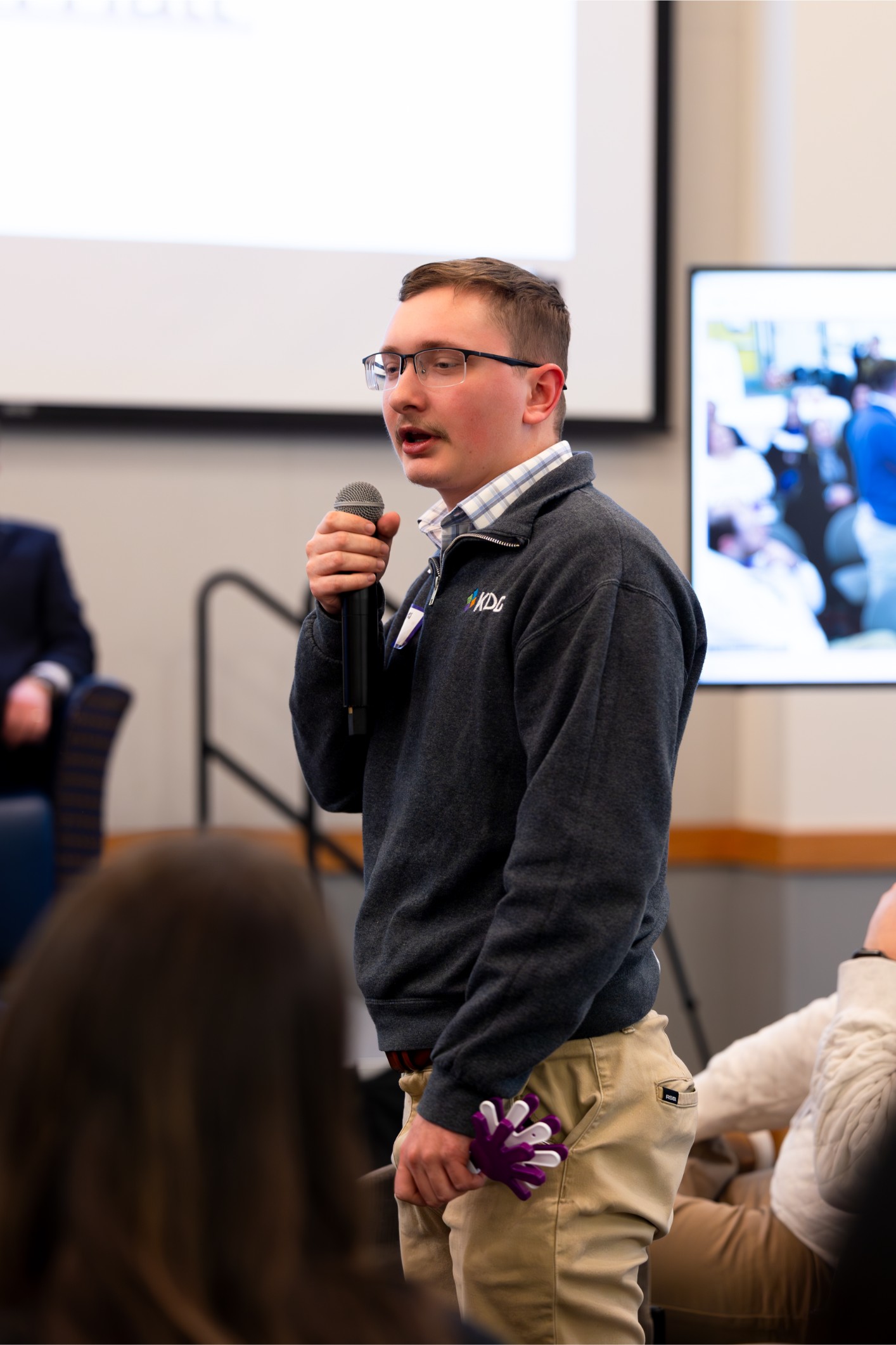 A person in a gray sweater holds a microphone while speaking at an indoor event. A person in a gray sweater holds a microphone while speaking at an indoor event.