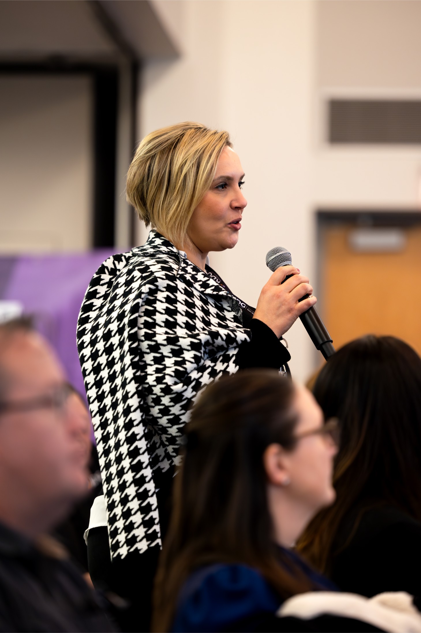 A person wearing a black and white houndstooth jacket speaks into a microphone, standing among seated audience members. A person wearing a black and white houndstooth jacket speaks into a microphone, standing among seated audience members.