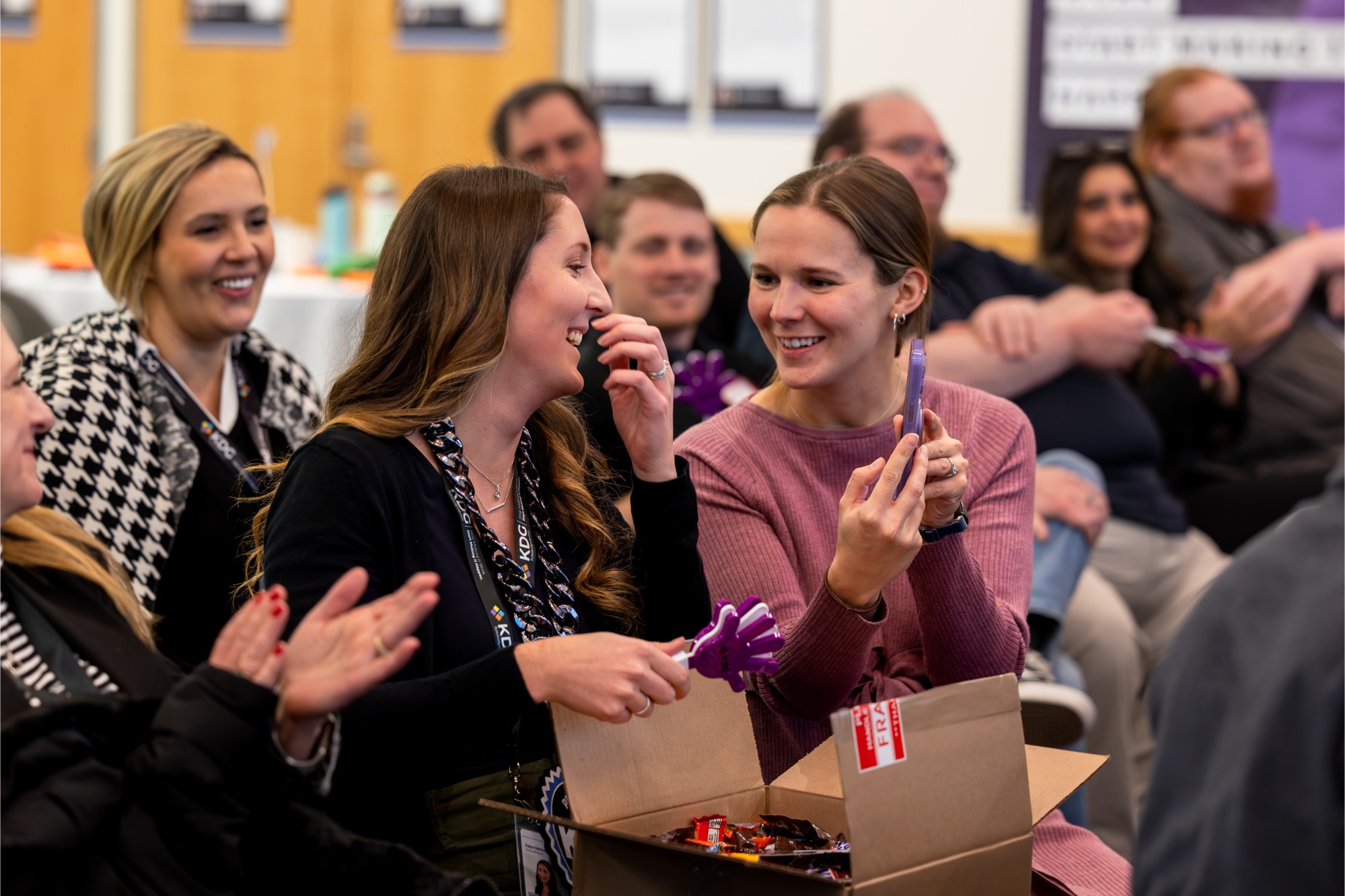 A group of people sitting and clapping, with two women in the foreground smiling and holding purple items from a box. A group of people sitting and clapping, with two women in the foreground smiling and holding purple items from a box.