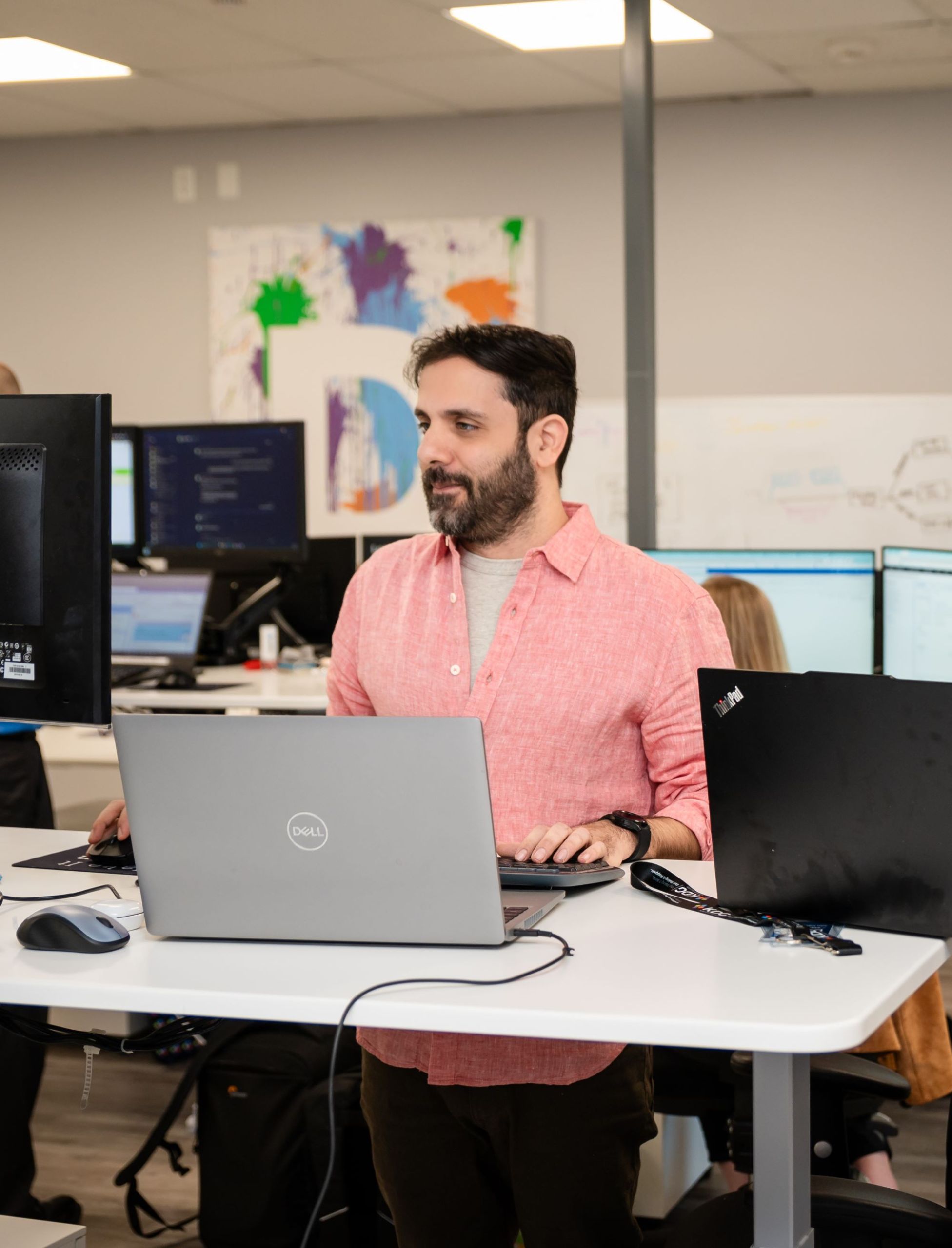 A man in a pink shirt diligently focuses at his standing desk, surrounded by laptops and monitors