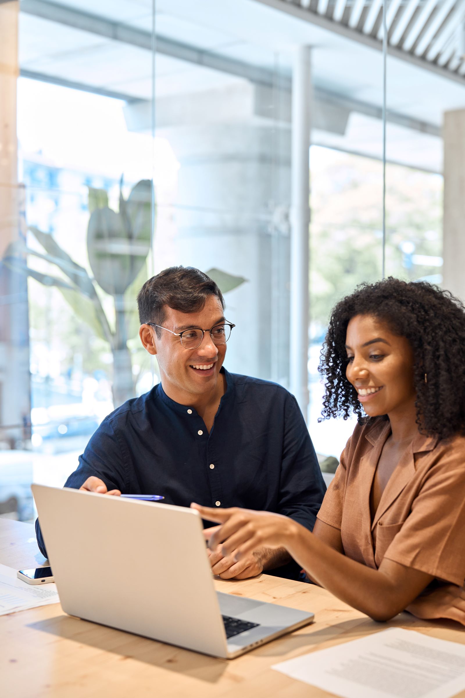 Two people sit at a table in a modern office, smiling and discussing something on a laptop. Papers and a pen are on the table as they review options for launching their new no code payment page.