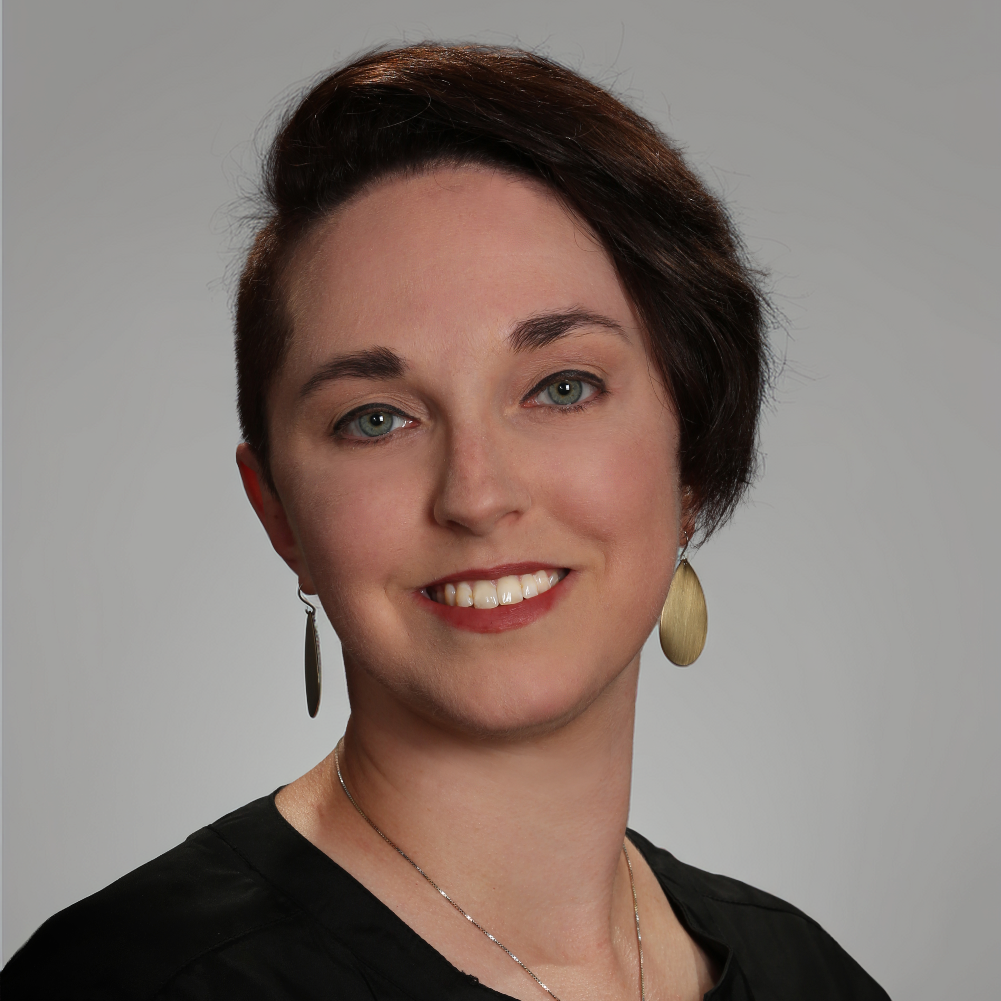 Caitlin Nelson, with short dark hair styled to one side and striking green eyes, wears oval earrings, a black top, and a necklace, smiling at the camera against a plain gray background. Caitlin Nelson, with short dark hair styled to one side and striking green eyes, wears oval earrings, a black top, and a necklace, smiling at the camera against a plain gray background.