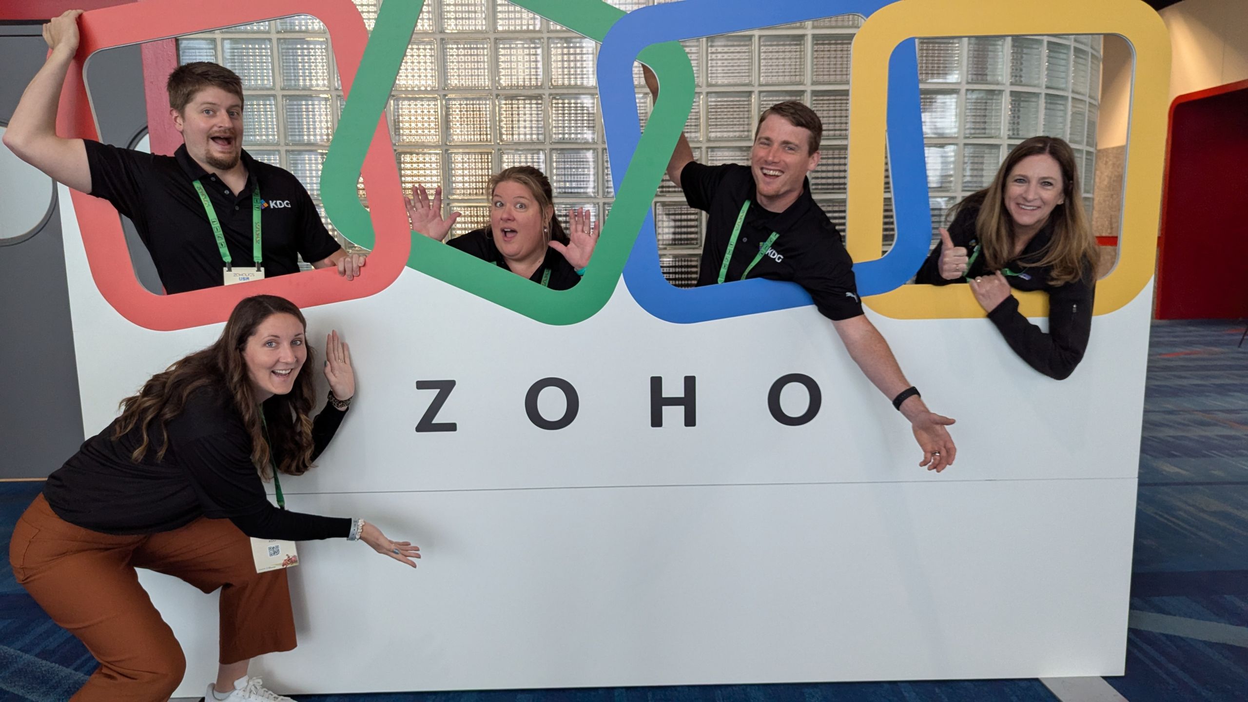 Five members of the KDG leadership team pose playfully with a large Zoho sign featuring colorful frames, inside a convention center at Zoholics 2025.