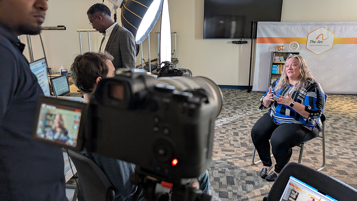 A woman from The Arc of Lehigh & Northampton Counties sits and speaks in a recorded interview by Zoho Creator and KDG teams setup with cameras, crew, and studio lighting.
