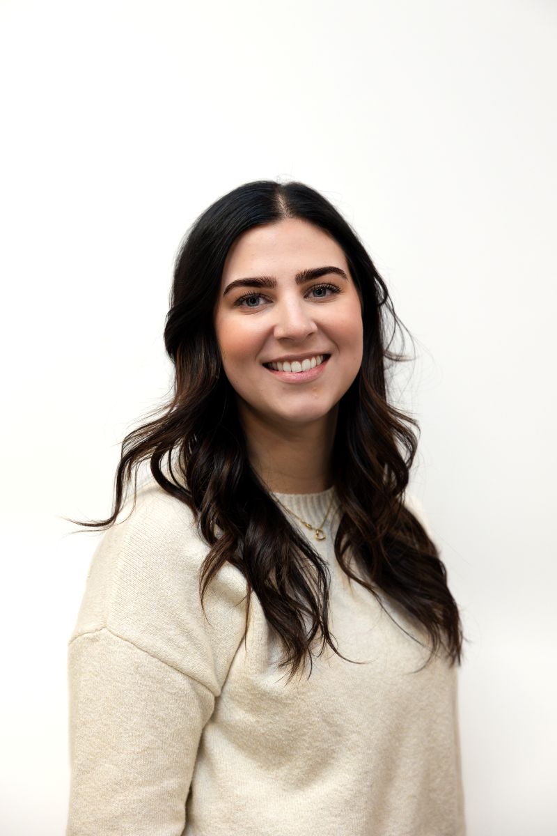 A woman with long dark hair, wearing a cream-colored sweater, smiles in front of a plain white background. A woman with long dark hair, wearing a cream-colored sweater, smiles in front of a plain white background.