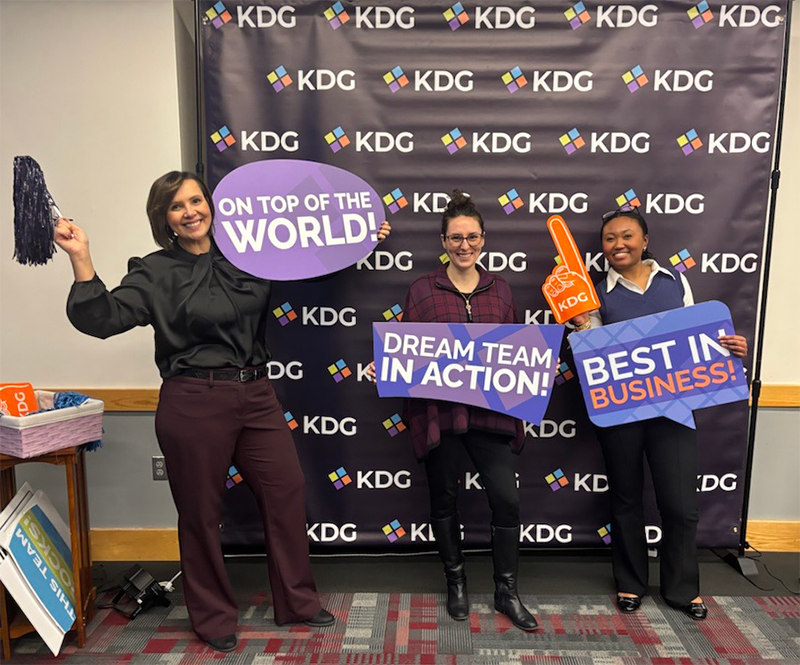 Three people stand in front of a KDG-branded backdrop, holding signs that say "ON TOP OF THE WORLD!", "DREAM TEAM IN ACTION!", and "BEST IN PROFESSIONAL SERVICES BUSINESS PROCESS!.