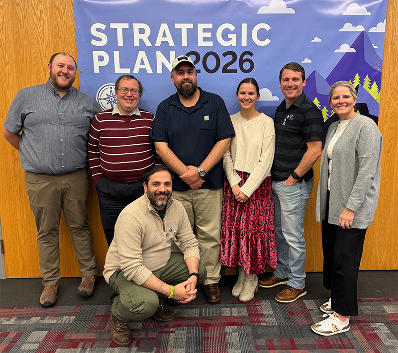 Seven people stand side by side in front of a "Strategic Plan 2026" banner, representing the Professional Services team, posing for a group photo indoors on a patterned carpet.