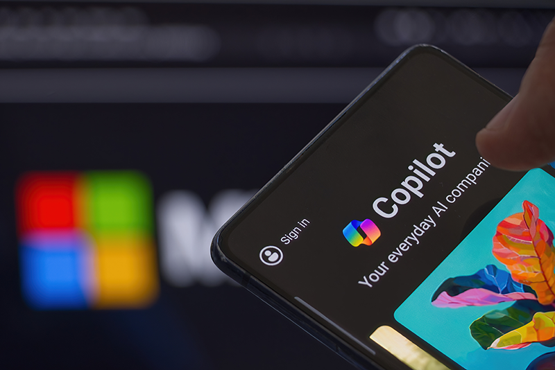 A smartphone screen displays the Microsoft Copilot app interface, featuring a sign-in option and colorful logo, with seamless integration for Bing Search, in front of a blurred Microsoft logo.